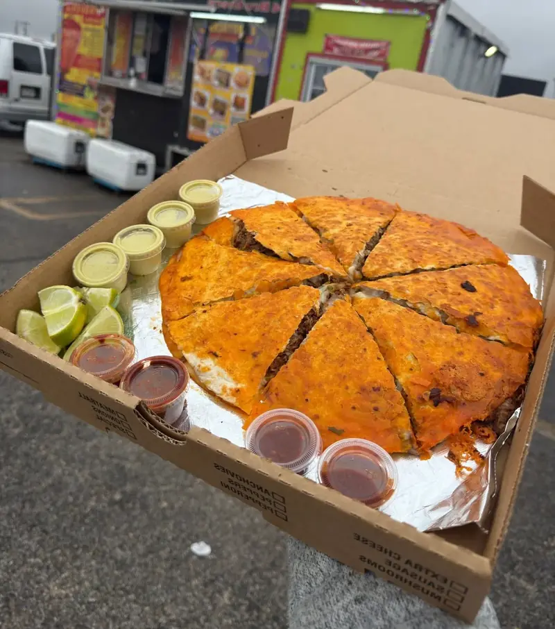 Pizza Birria in a takeout box with limes and salsa cups, food truck in the background